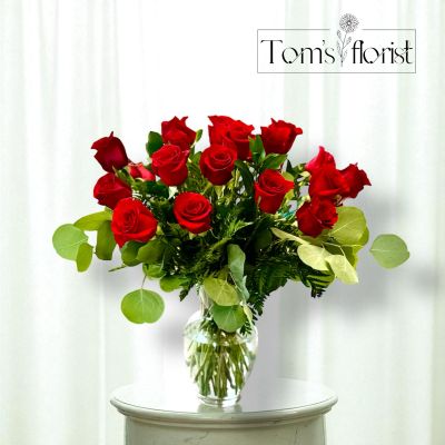 Tom's Florist Smyrna - The image features a classic and elegant floral arrangement of red roses in a clear glass vase. The bouquet is composed of two dozen vibrant red roses, beautifully arranged with lush green foliage, including eucalyptus and ferns. The deep red color of the roses contrasts beautifully with the rich green leaves, creating a timeless and romantic display. This arrangement is perfect for expressing love, admiration, or celebrating a special occasion.