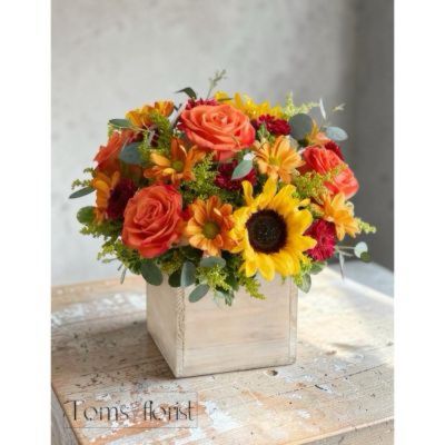 Tom's Florist Smyrna - EXCLUSIVE The color and charm of an autumn country harvest inspired our farmhouse-style bouquet. A loose, natural gathering of blooms in vibrant shades of red, orange and yellow creates the perfect complement to our rustic, grey-washed wooden cube. With its soft tones and natural textures, it’s an ideal container for flowers and plants, adding warm and coziness to any fall décor.
