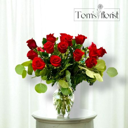 Tom's Florist Smyrna - The image features a classic and elegant floral arrangement of red roses in a clear glass vase. The bouquet is composed of two dozen vibrant red roses, beautifully arranged with lush green foliage, including eucalyptus and ferns. The deep red color of the roses contrasts beautifully with the rich green leaves, creating a timeless and romantic display. This arrangement is perfect for expressing love, admiration, or celebrating a special occasion.