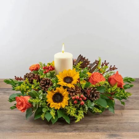 Tom's Florist Smyrna - This fall-themed centerpiece features vibrant sunflowers paired with rich, warm-toned roses, a shade of deep orange.  Accented with pine, and sprigs of greenery, it captures the essence of the season's cozy, harvest-inspired charm. Perfect for a festive table or autumn celebration.