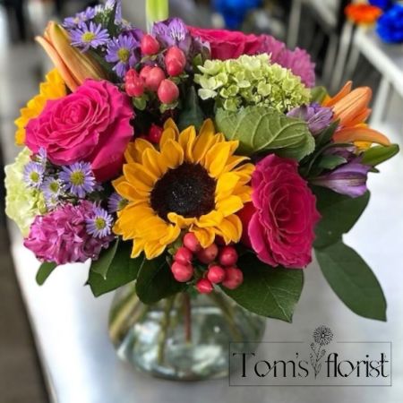 Tom's Florist Smyrna - Experience the warmth and joy of a perfect sunset with Sunset Bliss, a striking floral arrangement that celebrates the beauty of vibrant color. Bursting with shades of hot pink, purple, and yellow, this lively mix of sunflowers, roses, and lilies creates a radiant display that evokes the brilliance of a summer evening sky. The bold blooms come together in a symphony of color, radiating warmth and energy to brighten any space.

Whether you're looking to bring a cheerful touch to your home or gift someone a burst of sunshine, Sunset Bliss is the perfect way to add a lively, colorful accent. Let this arrangement fill your world with the glow of endless sunsets and boundless joy.