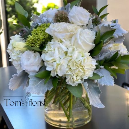 Tom's Florist Smyrna - Indulge in the serene beauty of Timeless Elegance, a sophisticated floral arrangement that radiates purity and grace. This exquisite design combines the soft, ethereal glow of white hydrangeas with the classic charm of white roses, all nestled together in a harmonious blend of delicate textures. The addition of lush green accents adds a subtle touch of natural vibrance, creating an arrangement that speaks to luxury and refined taste.

Whether as a thoughtful gift or a refined addition to your space, Timeless Elegance is the embodiment of opulence and understated beauty, leaving a lasting impression with every petal.

