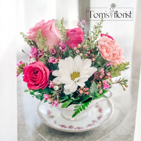 Tom's Florist Smyrna - Step into a world of elegance and charm with Tea Time Charm, a beautifully arranged floral creation presented in a vintage porcelain tea cup and matching saucer. This delicate arrangement is perfect for celebrating life’s sweetest moments, bringing a touch of tea party sophistication to any space.

Soft and graceful, this petite bouquet features lush pink roses, a fragrant pink carnation, cheerful white daisies, and delicate pink wax flowers, all accented with golden solidago for a warm, whimsical touch. Whether given as a thoughtful gift or used as a charming centerpiece, this timeless design exudes grace and beauty.

Perfect For:
- Tea party lovers and vintage enthusiasts

- Birthday surprises, get-well wishes, or just because

- Mother’s Day, bridal showers, or special celebrations

- Adding a touch of floral elegance to any desk or table

Sip in the beauty of Tea Time Charm, a floral arrangement that blends classic elegance with fresh floral delight—the perfect gift for anyone who loves tea, flowers, and timeless beauty.

Teacup and saucer set may vary based on availability. 

