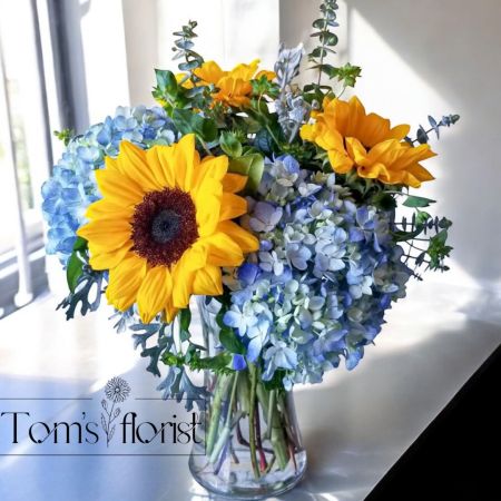 Tom's Florist Smyrna - Brighten any occasion with the cheerful charm of Sunflower Skies. This stunning arrangement combines the vibrant yellow of sunflowers with the calming blue of premium hydrangeas, creating a beautiful contrast that radiates warmth and happiness. Perfect for birthdays, get well wishes, or simply as a thoughtful gesture, Sunflower Skies brings a touch of sunshine and elegance to any moment. With its lively yet refined style, this arrangement is a timeless choice for spreading joy, love, and good spirits.

